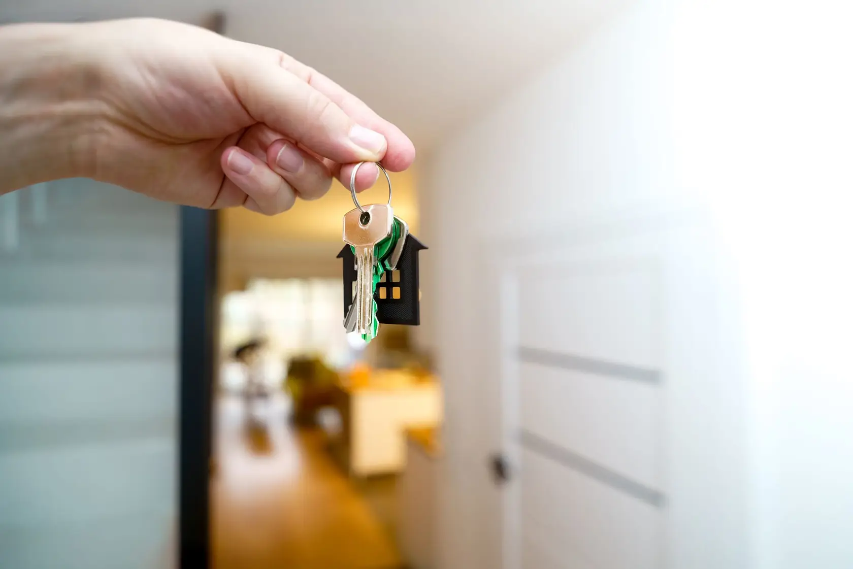 Main tenant un trousseau de clés avec un porte-clés en forme de maison, devant l’entrée d’un appartement