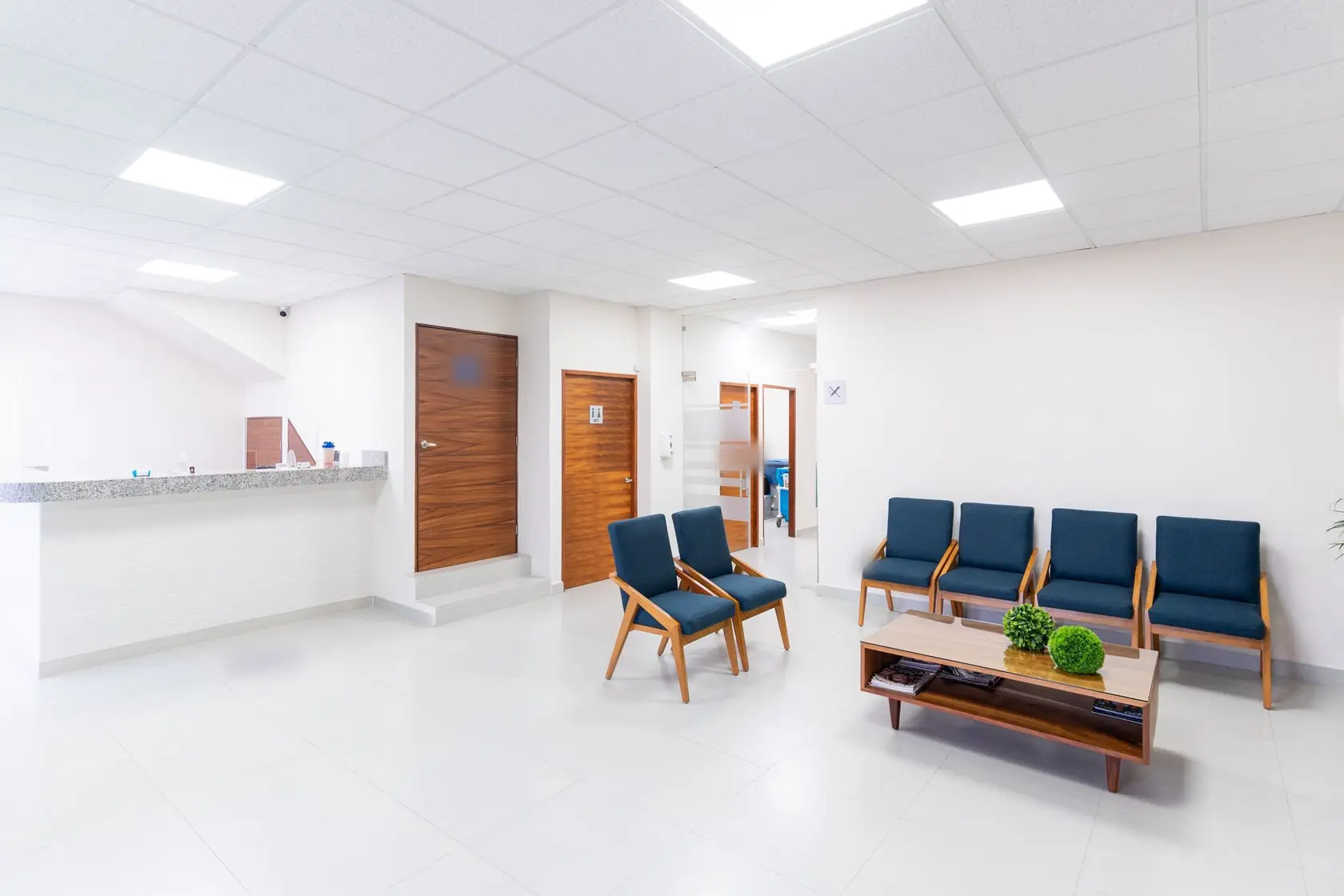 Salle d’attente claire : fauteuils bleus autour d’une table basse, banque d’accueil et portes en bois au fond