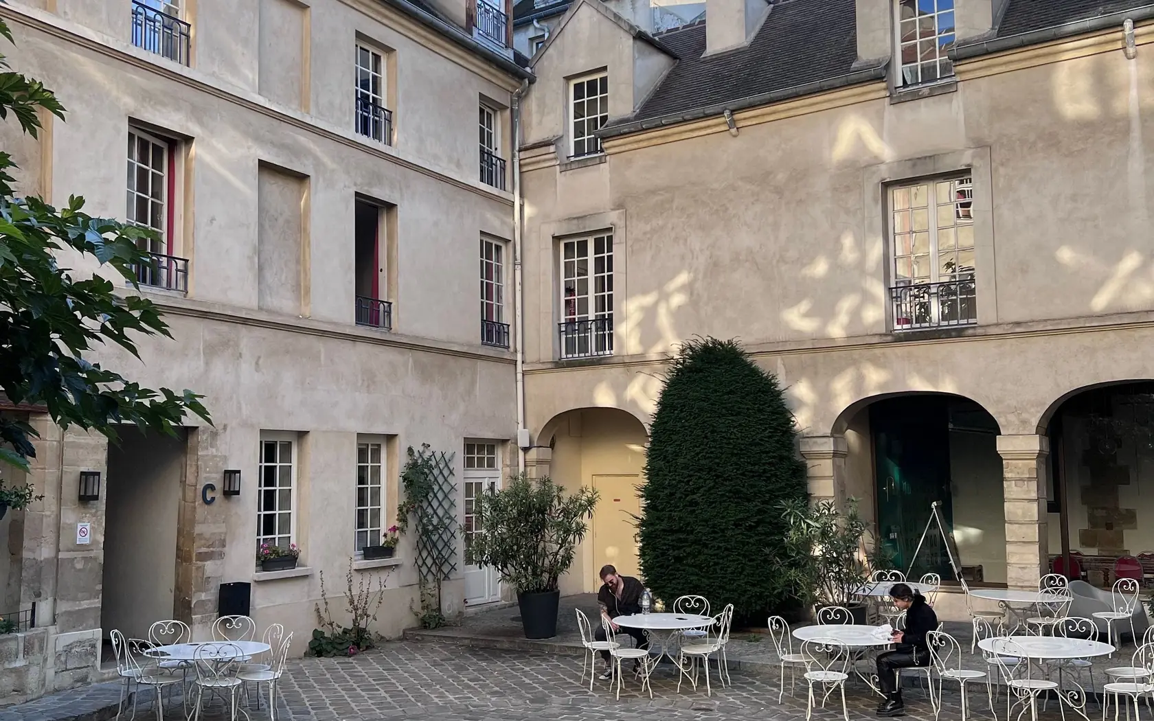 Cour pavée d’un immeuble ancien aux façades claires, avec tables de café et plantes en pot