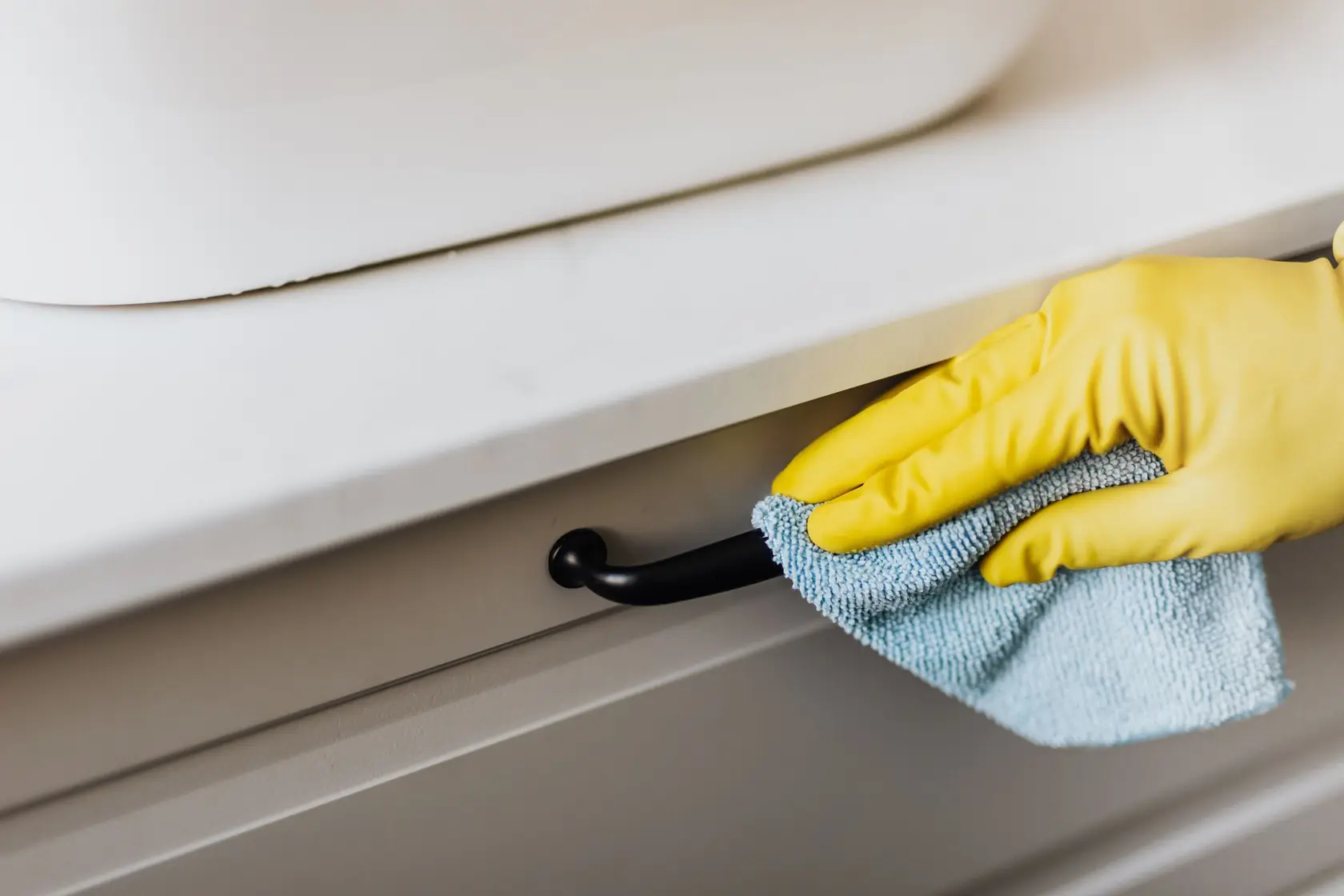 Main en gant jaune essuyant la poignée d’un tiroir avec une microfibre bleue
