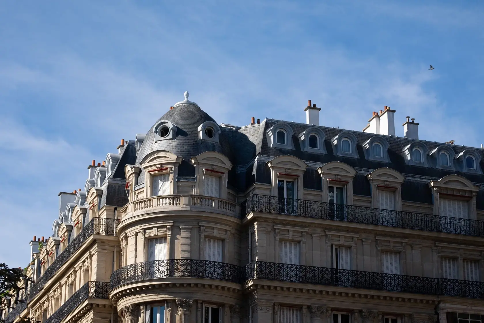 Façade d’immeuble haussmannien en pierre de taille, balcons filants en fer forgé et toit d’ardoise sous un ciel bleu