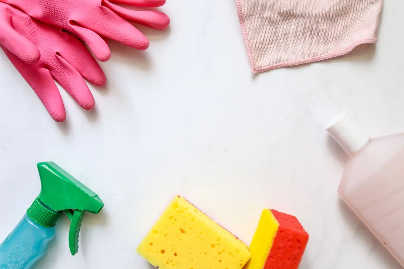 Gants de ménage roses, chiffon microfibre, éponges et pulvérisateur posés sur un plan blanc