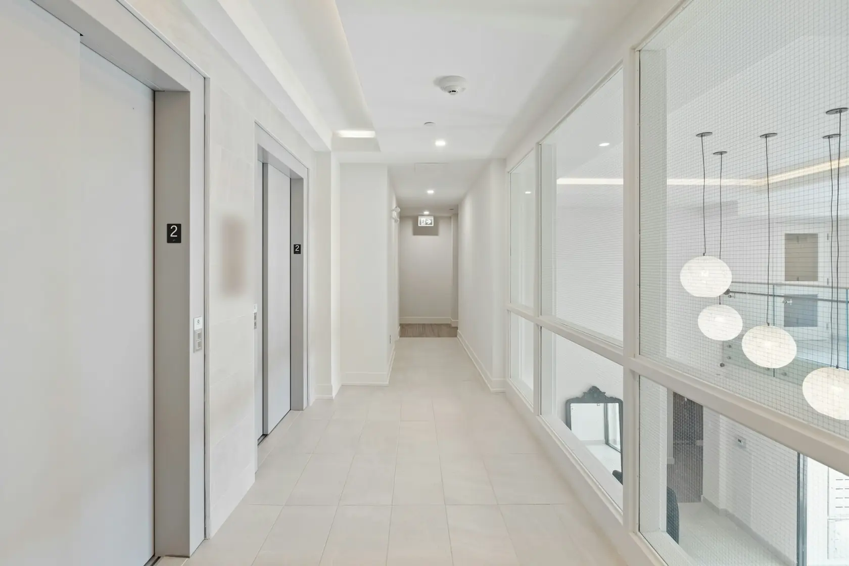 Couloir d’étage clair aux portes d’ascenseur métalliques, bordé d’une paroi vitrée et de suspensions en verre
